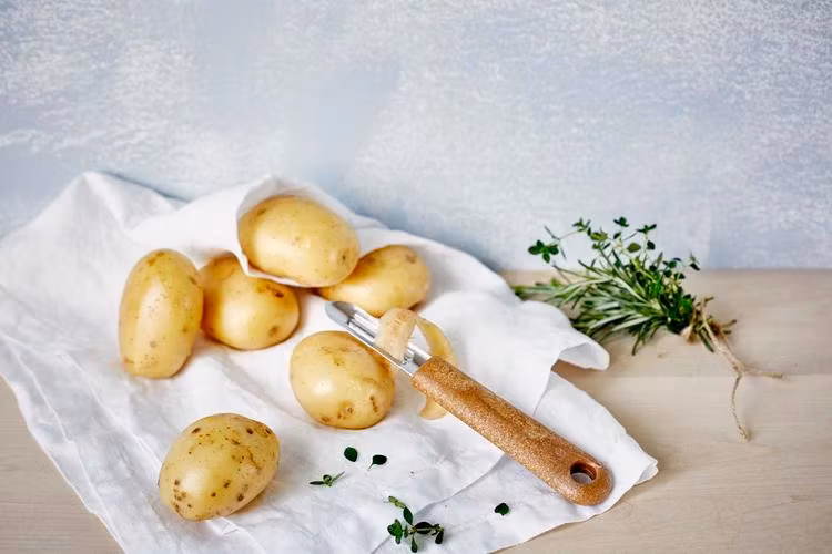 Naturfärgad potatisskalare i rostfritt stål med skaft av bioplast och träfiber på en vit duk med potatis brevid.