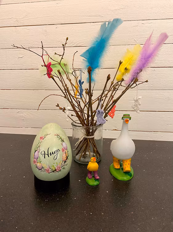 Grönt påskägg med blom- och äggkransmotiv bredvid påskris och prydnadsfigurer