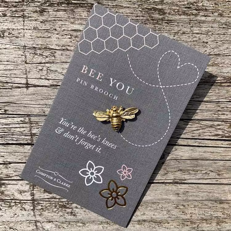 Bild på en pin i form av ett bi tillverkat i tenn som guldpläterats. Pinen är fäst på ett litet kort med texten "bee you"