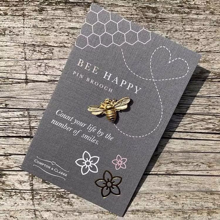 Bild på en pin i form av ett bi tillverkat i tenn som guldpläterats. Pinen är fäst på ett litet kort med texten "bee happy"