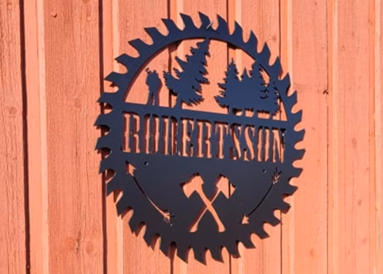Svart skylt med eget namn skuren i metall, mot röd trävägg.
