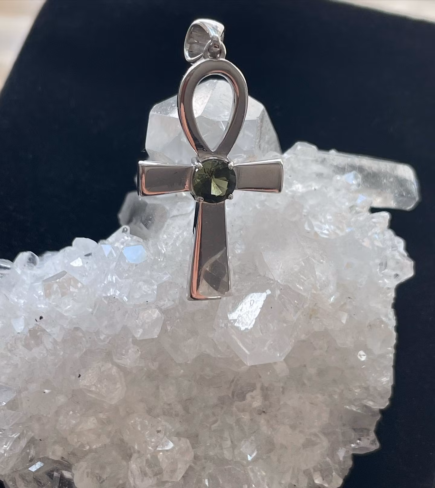 Bilden visar ett silversmycke med en Ankh symbol i silver med en grön Moldavit i mitten.