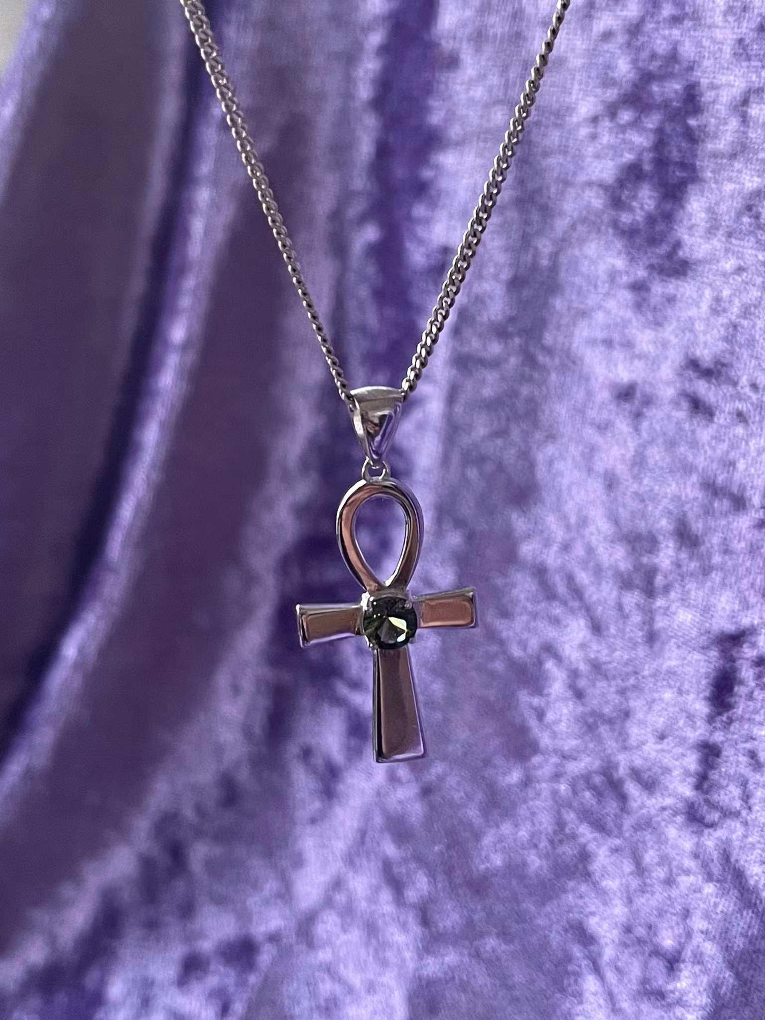 Bilden visar ett silversmycke med en Ankh symbol i silver med en grön Moldavit i mitten.