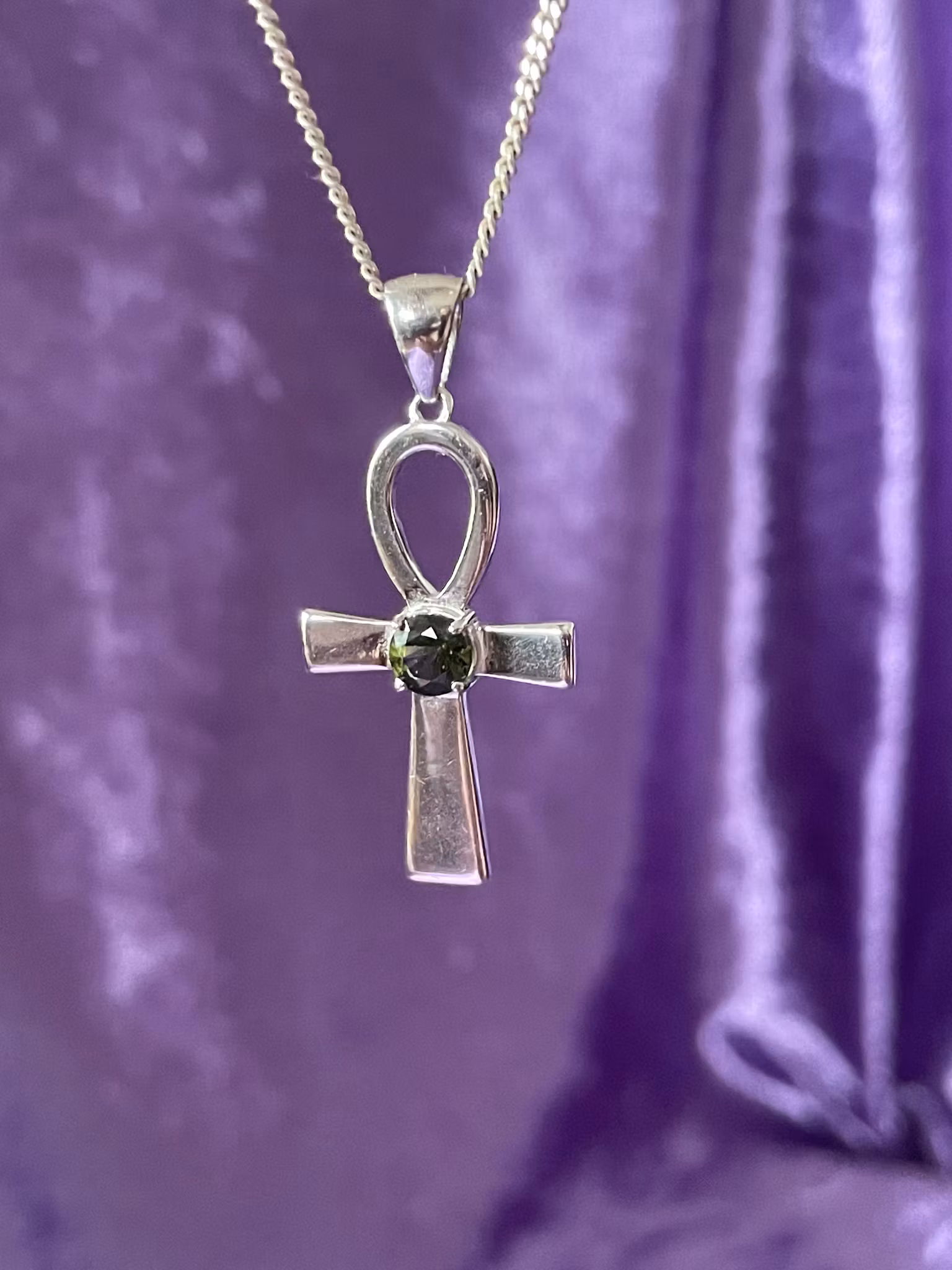 Bilden visar ett silversmycke med en Ankh symbol i silver med en grön Moldavit i mitten.