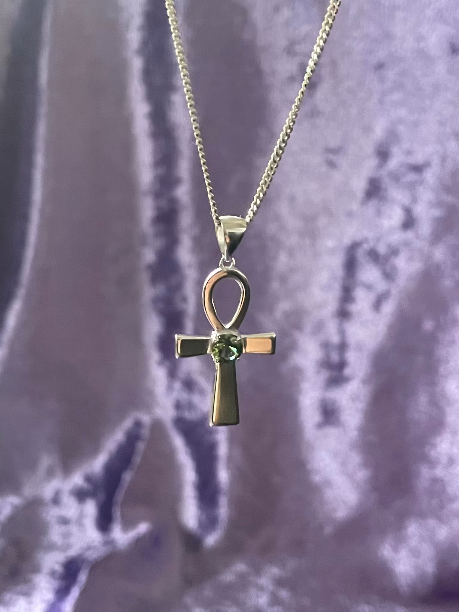 Bilden visar ett silversmycke med en Ankh symbol i silver med en grön Moldavit i mitten.