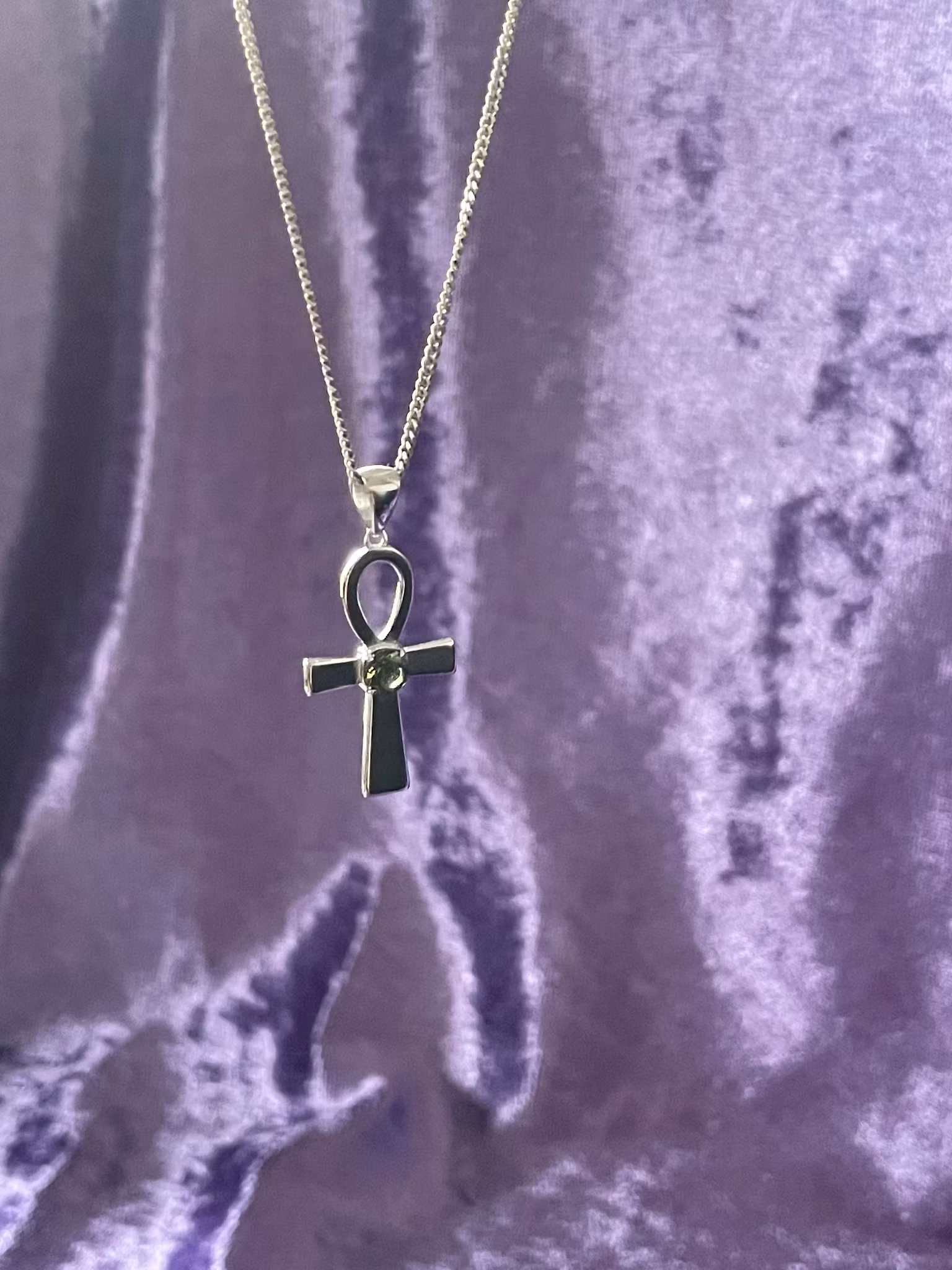 Bilden visar ett silversmycke med en Ankh symbol i silver med en grön Moldavit i mitten.