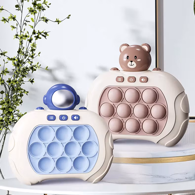 Två interaktiva leksaker med pop-it-knappar, en med en blå astronautfigur och en med ett brunt björnhuvud. Båda har knappar för ljud och ljus och en färgglad, bubblig design. De står på en vit yta.