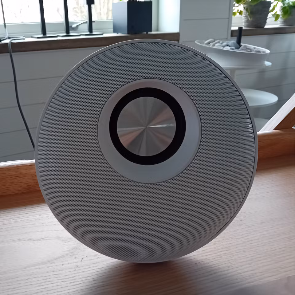 En rund, modern högtalare med metalliskt galleri och en central detalj i borstat stål, placerad på ett träbord i ett ljust rum