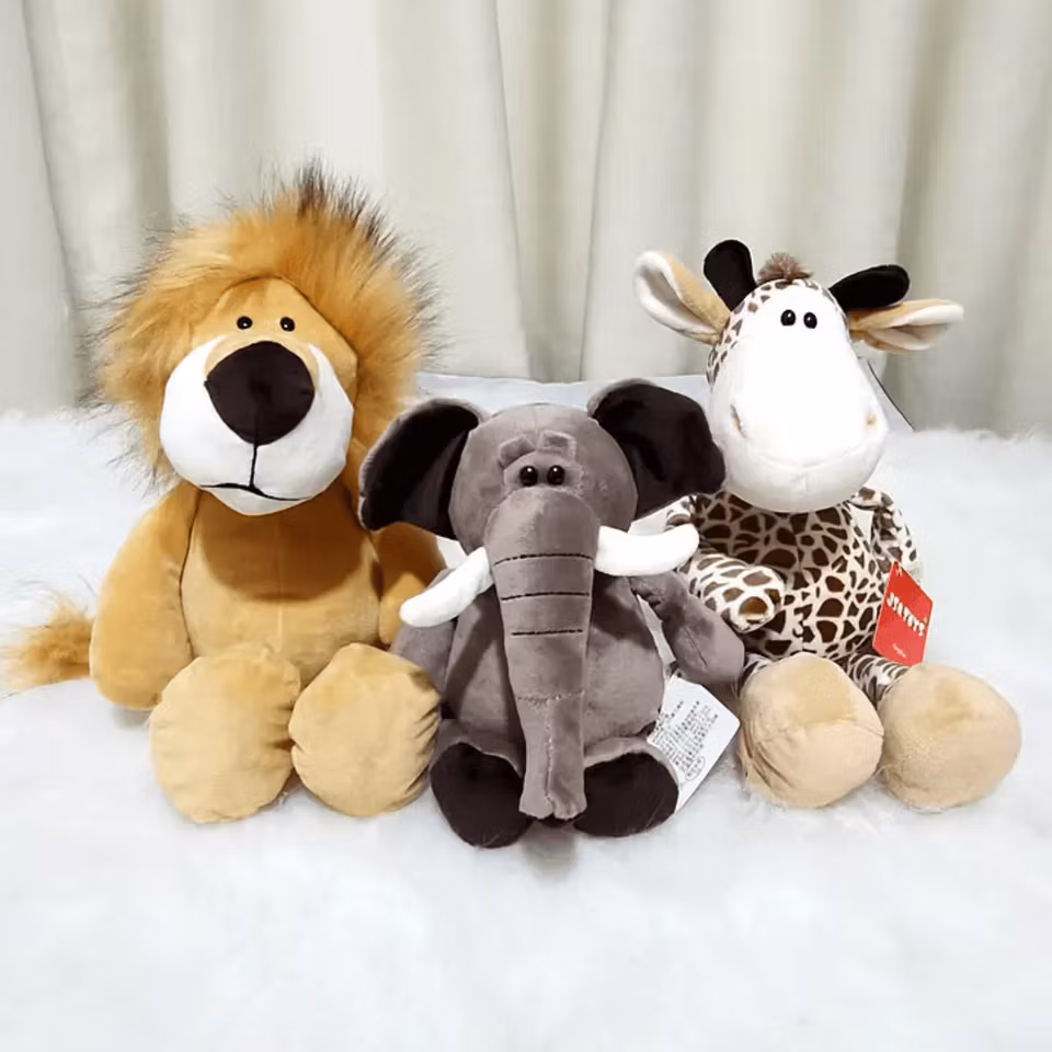 Tre gosedjur föreställande en lejon, en elefant och en giraff sitter bredvid varandra på en vit, fluffig yta med en ljus gardin i bakgrunden. Lejonet har en gulbrun kropp och en fluffig man.