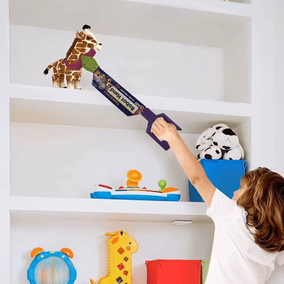 Ett barn i vit tröja använder en leksaksgrepparm för att nå en gosedjursgiraff på en hög hylla i ett vitt förvaringsskåp fyllt med leksaker och mjukisdjur