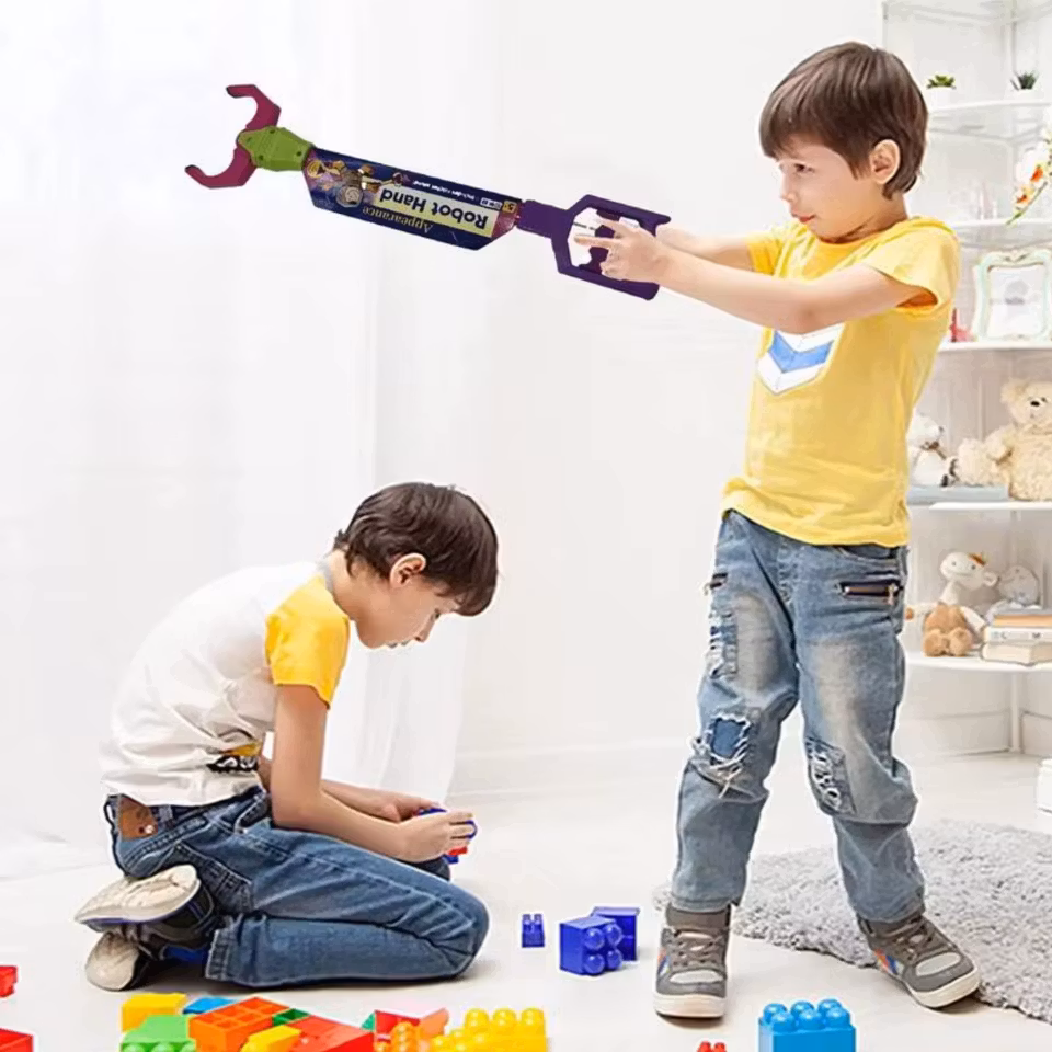 Två barn leker i ett ljust rum. Ett barn i gul tröja håller en leksakstänger med texten 'Robot Hand' och riktar mot det andra barnet, som sitter på golvet och bygger med färgglada klossar.