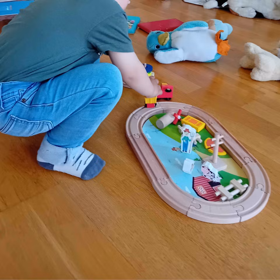 Träleksak i bondgårdstema med en oval tågbana, röd traktor med gul släpvagn, en lada, staket, vindmölla och figurer av en bonde, ko och häst. Bakgrunden visar en tecknad bondgård med djur och en bonde