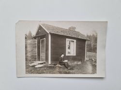 Vykort bild på person som läser bok framför en stuga.
