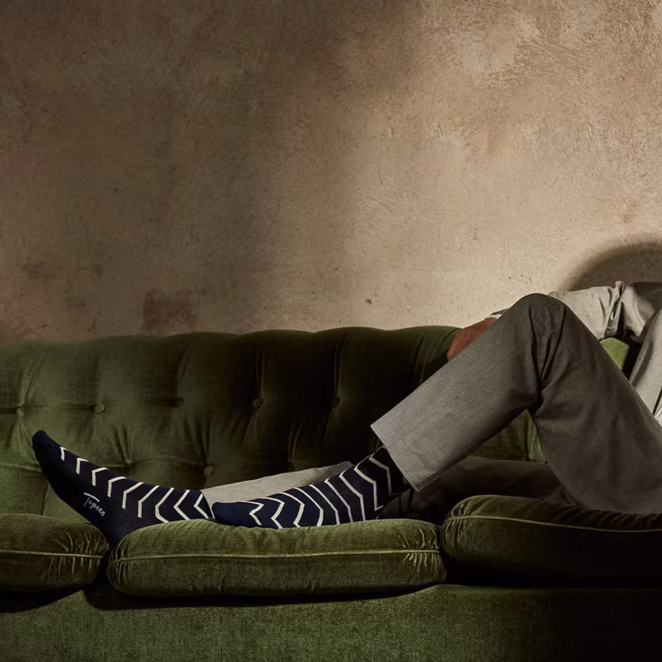 Man sitter avslappnat på soffa med båda fötterna uppe, bär TOPECO Sock Zigge herrstrumpor i bambu marinblå
