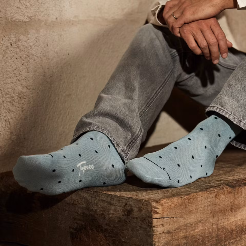 Man sitter på soffa och kramar knäna, bär TOPECO Sock Barslöv herrstrumpor i bambu vintagegrön med prickmönster, strumporna synliga