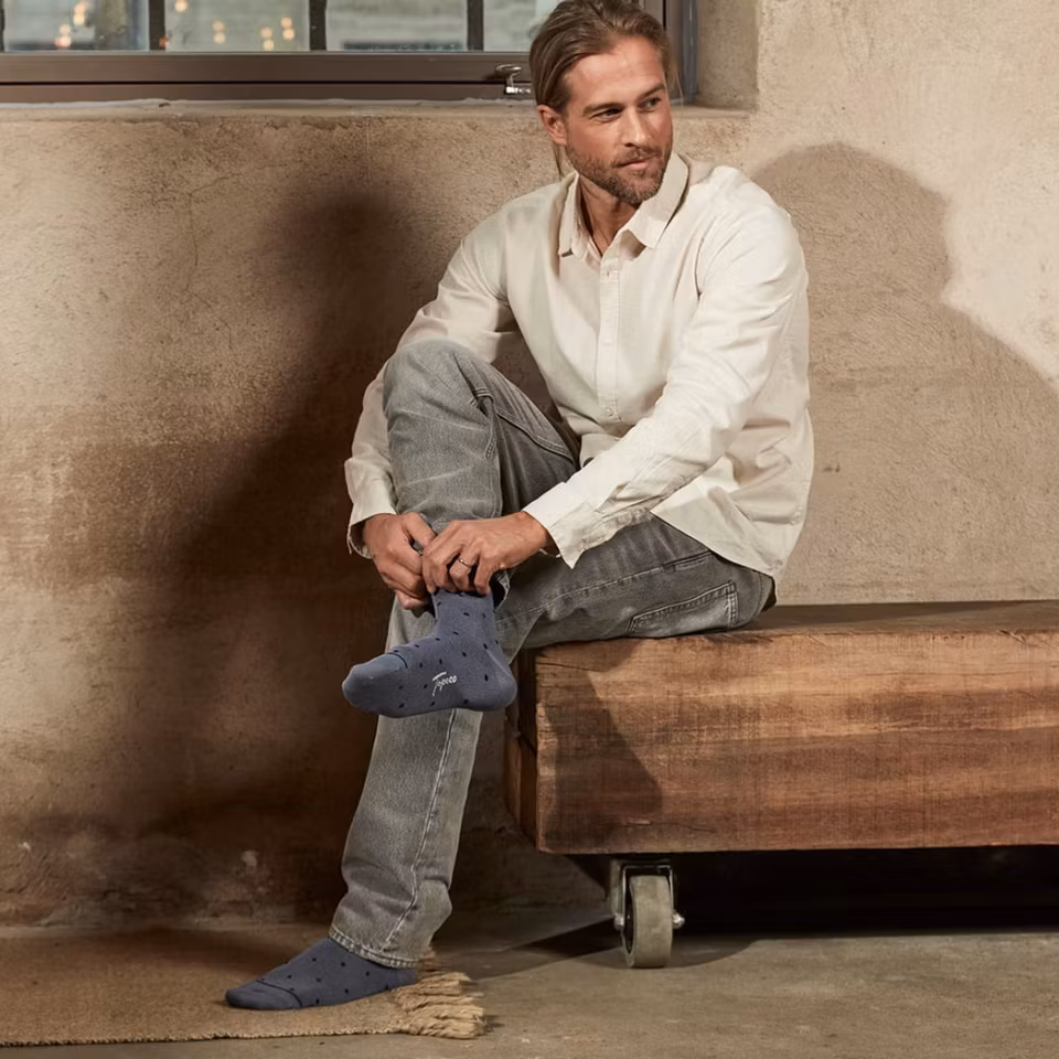 Man sitter på en träbänk i ljus skjorta och grå jeans, bär TOPECO Sock Bårslöv herrstrumpor i bambu ljusblå med prickmönster