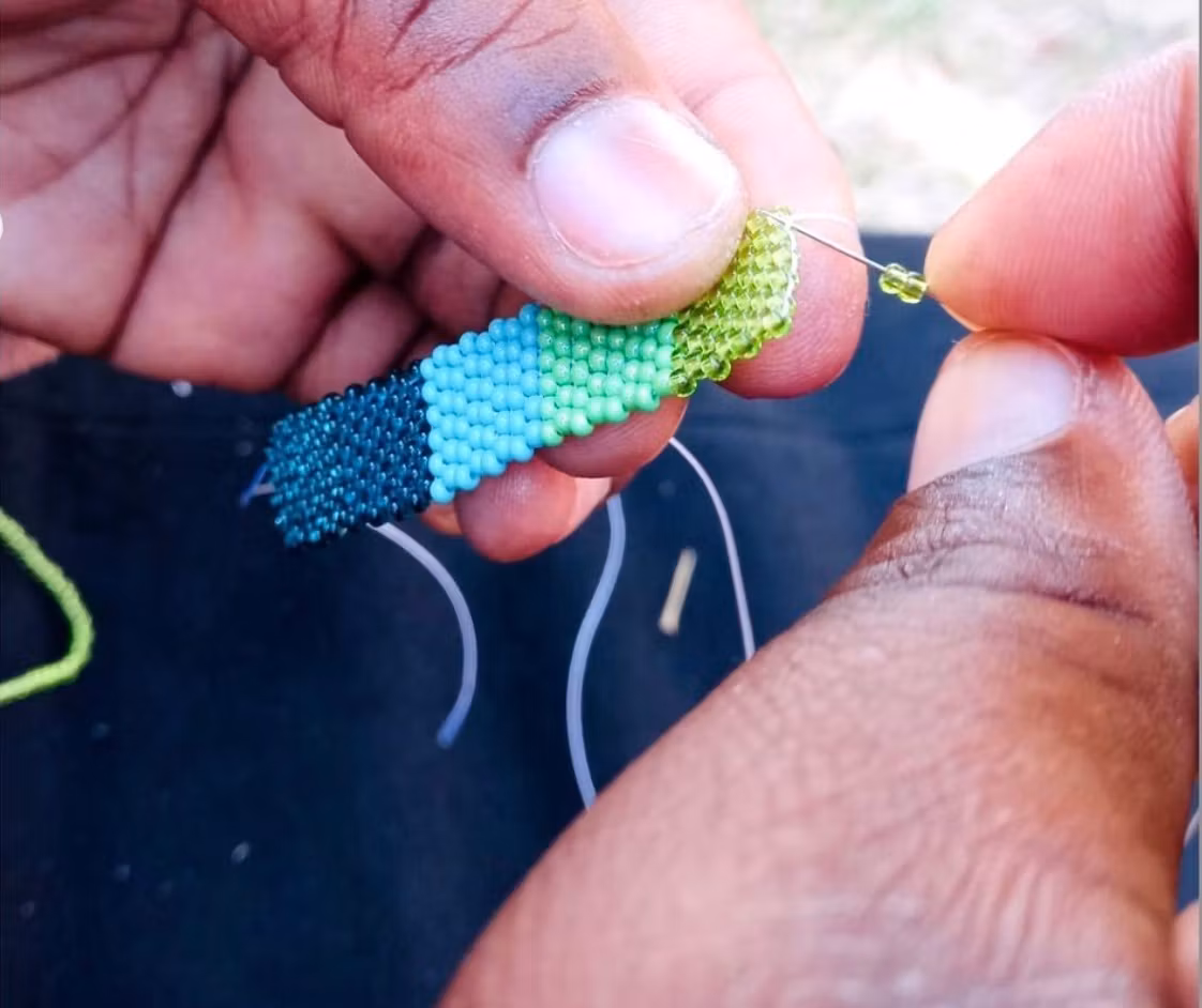 Armband av små glaspärlor i gröna nyanser. Bild där du ser hur pärlorna träs på tråden.Noggrant trådarbete, traditionellt Zulu-arbete i modern design. Sydafrika.