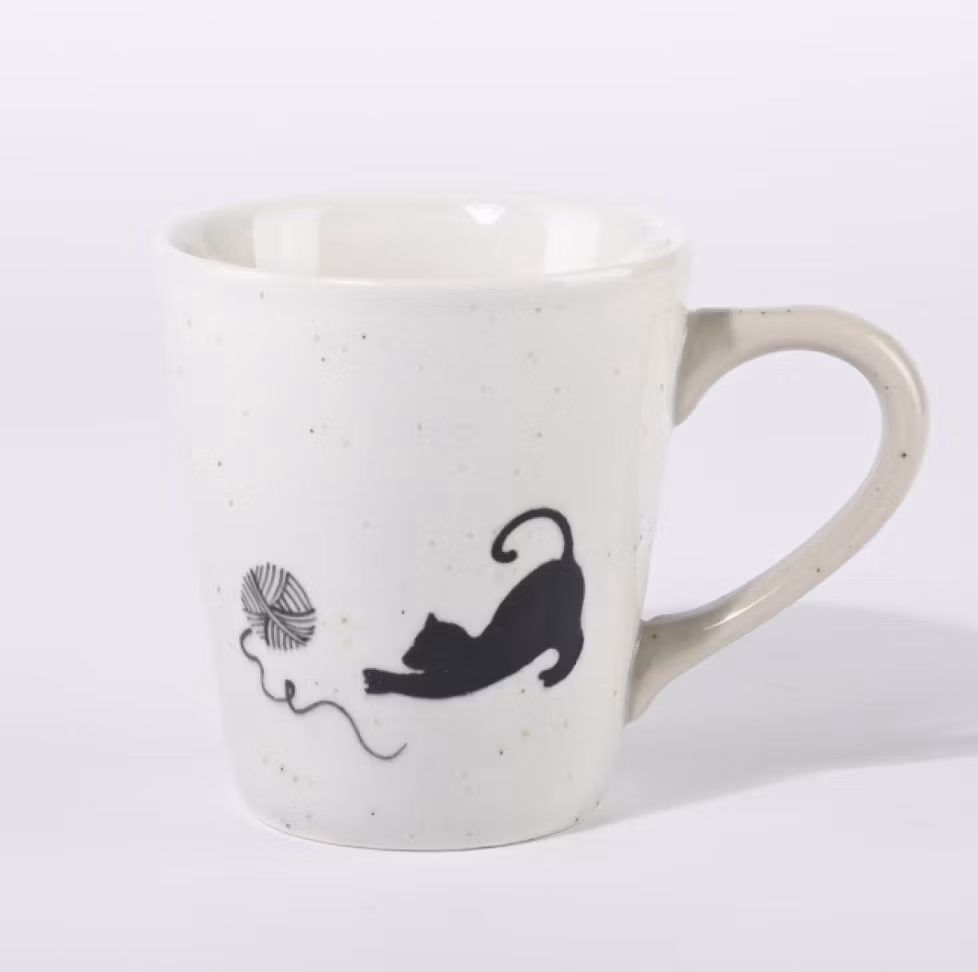 En beige-vit kopp till kaffe, te eller varm choklad. Motivet är en liten katt som leker med ett garnnystan, keramik. Fair Trade.