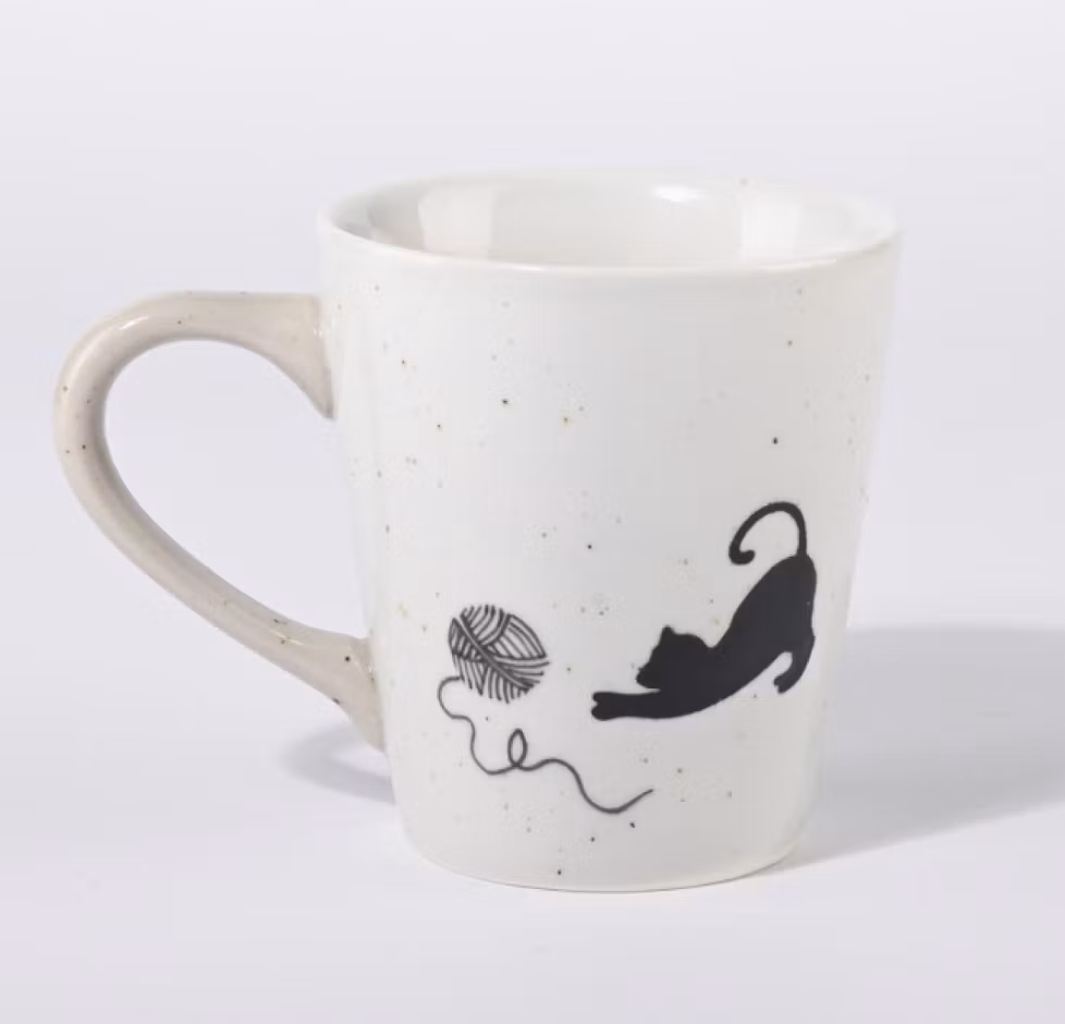 En beige-vit kopp till kaffe, te eller varm choklad. Motivet är en liten svart katt som leker med ett garnnystan, keramik. Motivet finns på båda sidorna. Fair Trade.