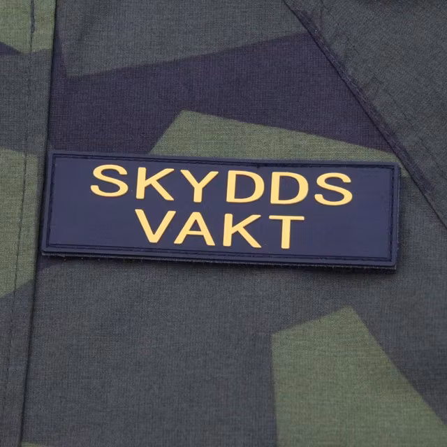 Ett Skyddsvakt PVC Avlång Kardborremärke med M90 kamouflage i bakgrunden.