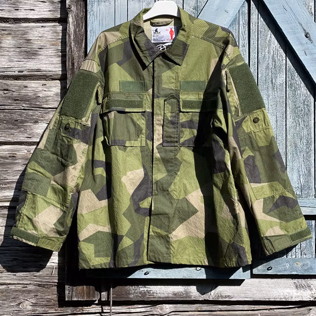 Seen full front is our popular Field Shirt M90 hanging on a wooded buildning as background