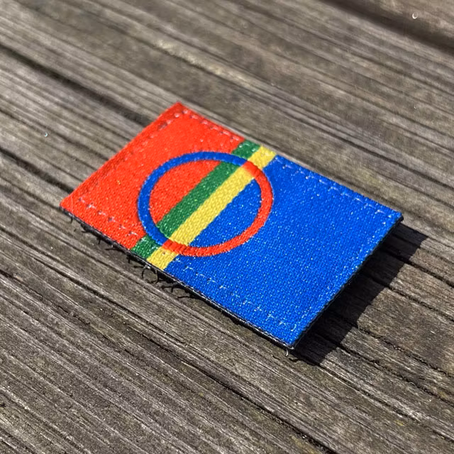 A Sámi Flag Hook Patch Small from TAC-UP GEAR laying flat on a wooded floor seen from an angle