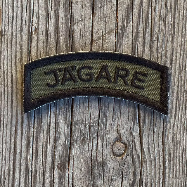 A JÄGARE K3 Type Hook Patch Black/Green seen from the front lying on wooded floor