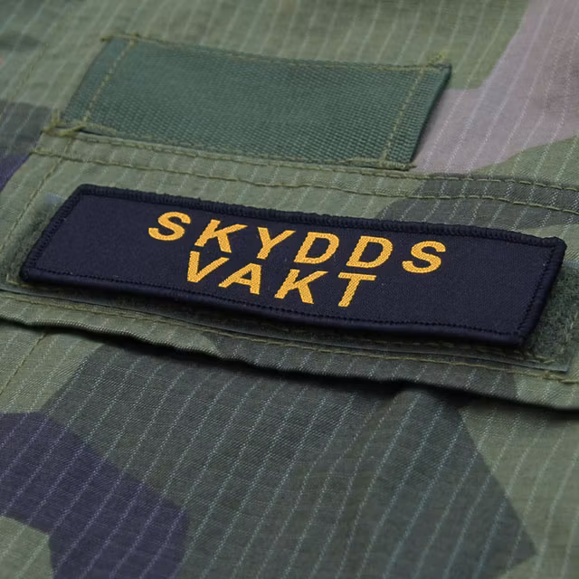 Skyddsvakt Avlång Kardborremärke med M90 kamouflage i bakgrunden.