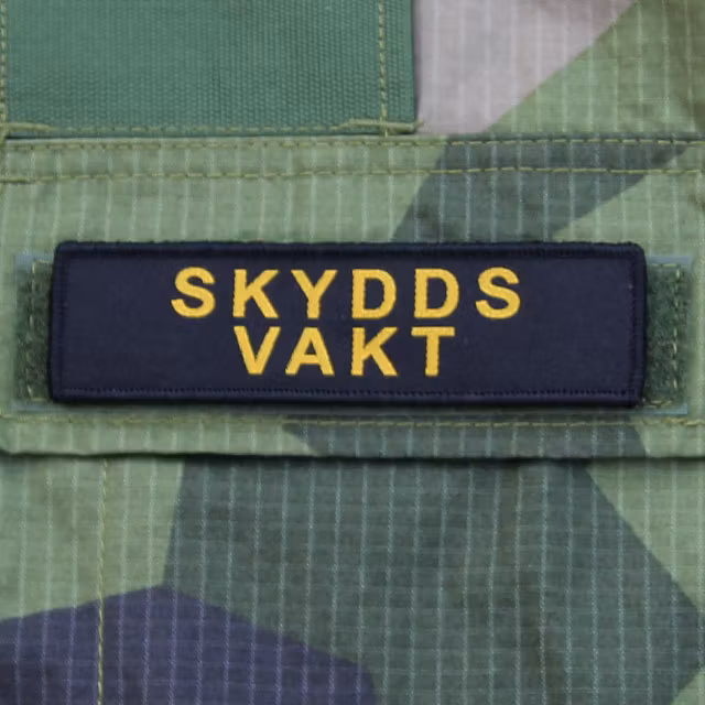 Ett Skyddsvakt Avlång Kardborremärke monterat på locket på en bröstficka på en M90 kamouflage jacka.