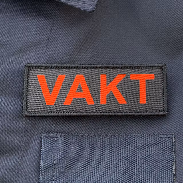 VAKT Röd text Kardborremärke mot blå jacka