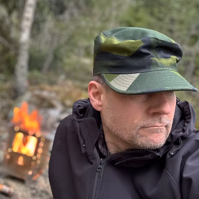 Patrol Cap M90 from TAC-UP GEAR on a model in the Swedish forest seen at a slight angle with a fire in the background