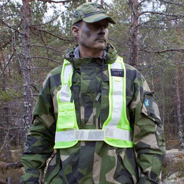 En Skyddsvakt Reflexsväst buren över en M90 kamouflage jacka och visar framsidan.