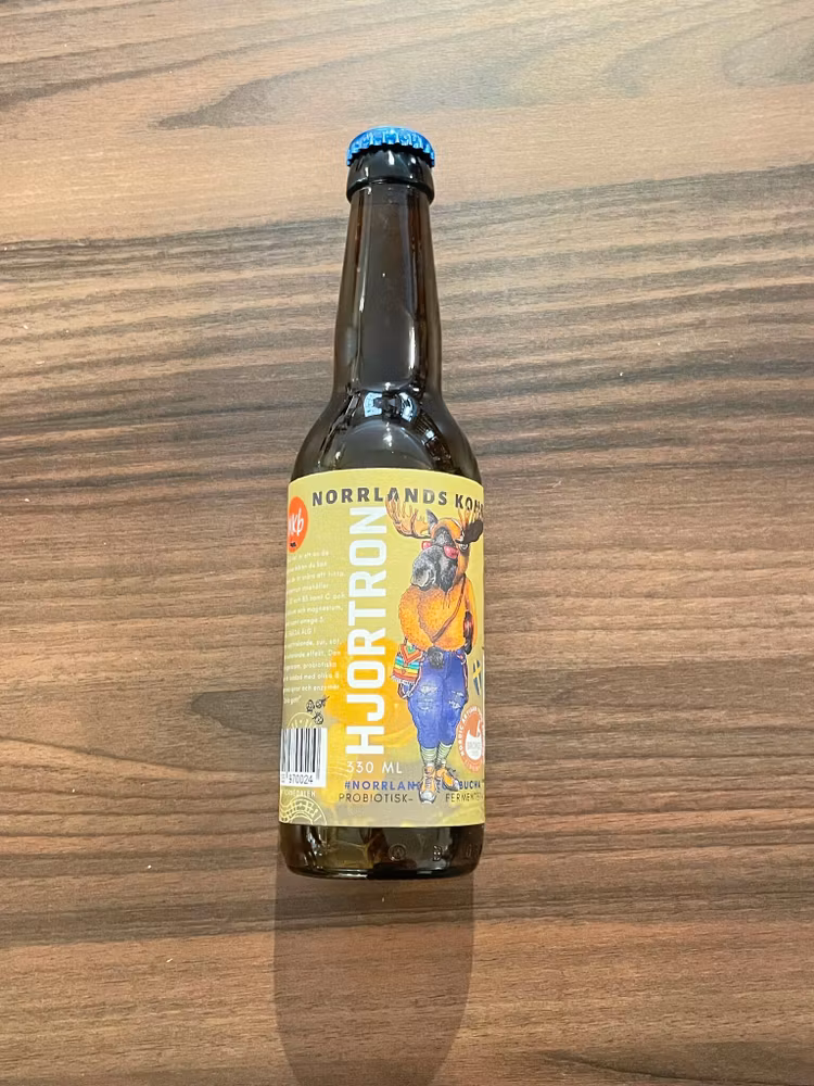 kombucha i brun glasflaska med gulgrön ettikett med en älg på. Smak av hjortron