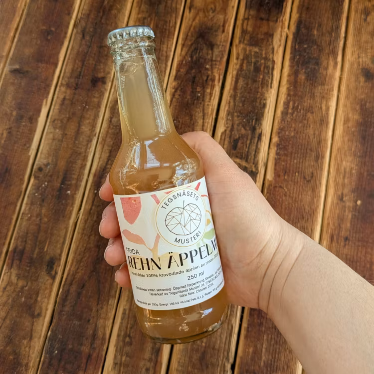 Äppelmust i flaska från Tegsnäsets musteri som hand håller i med träbakgrund