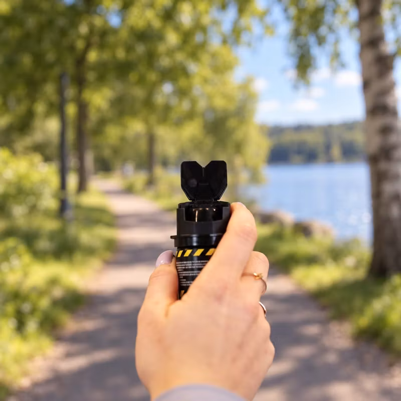Hand som håller en Bodyguard övningsspray med uppfällt lock som sikte, med en parkväg vid en sjö i bakgrunden